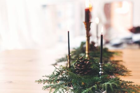 Stylish rustic christmas arrangement for festive dinner. Fir branches with pine cones and vintage candlestick with burning black candles on wooden table. Christmas rural table decor for feastの写真素材