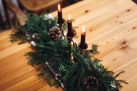 Fir branches with pine cones and vintage candlestick with burning black candles on wooden table. Stylish rustic christmas arrangement for festive dinner. Christmas rural table decor for feastの写真素材
