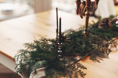 Stylish rustic christmas arrangement for festive dinner. Fir branches with pine cones and vintage candlestick with burning black candles on wooden table. Christmas rural table decor for feastの写真素材