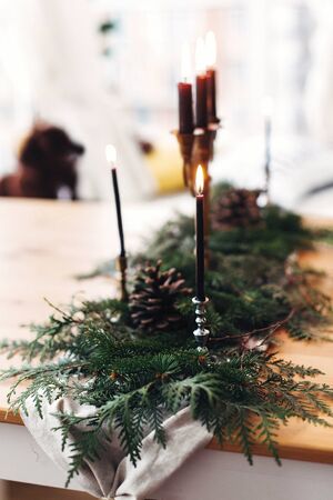 Stylish rustic christmas arrangement for festive dinner. Fir branches with pine cones and vintage candlestick with burning black candles on wooden table. Christmas rural table decor for feastの写真素材
