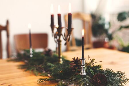Fir branches with pine cones and vintage candlestick with burning black candles on wooden table. Stylish rustic christmas arrangement for festive dinner. Christmas rural table decor for feastの写真素材