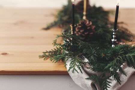 Stylish rustic christmas arrangement for festive dinner closeup. Fir branches with pine cones and vintage candlestick with black candles on linen cloth on wooden table. Christmas rural decorの写真素材