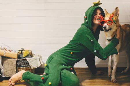 Happy girl in festive pajama hugging dog in holiday glasses with deer horns, sitting on floor with christmas presents in stylish room.の写真素材