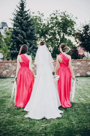 Gorgeous bride walking with bridesmaids in pink dresses, holding stylish bouquets in warm evening light in summer park. Beautiful happy bride with maid of honor posing togetherの写真素材