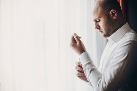 Groom putting on white shirt, standing near window. Stylish man getting ready in the morning in...