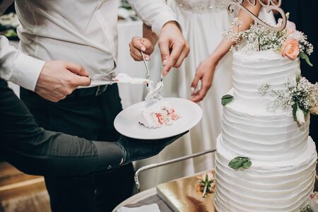 Bride and groom cutting stylish wedding cake at wedding reception in restaurant. Wedding couple holding slice of  wedding cake decorated with flowersの写真素材