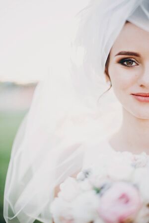 Sensual portrait of beautiful bride under veil holding bouquet outdoors. Gorgeous bride in white gown with modern wedding bouquet posing in summer park.の写真素材