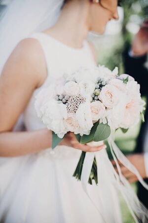 Modern wedding bouquet in bride hands. Gorgeous bride in white gown holding stylish bouquet of white and pink peonies and roses during walk outdoorsの写真素材