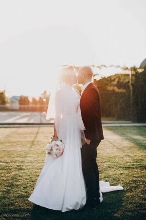 Stylish bride and groom gently kissing in sunset light. Beautiful bride with modern bouquet embracing with stylish groom in summer park. Sensual romantic momentの写真素材