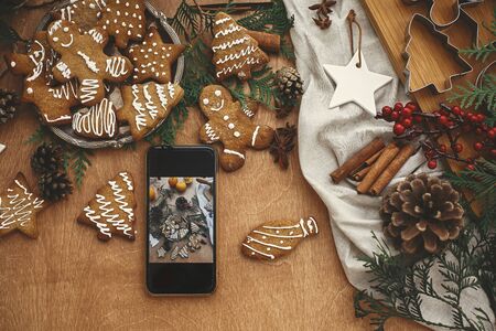 Phone with christmas photo of festive gingerbread cookies flat lay, anise, cinnamon, pine cones, cedar branches on rustic table. Merry Christmas. Holiday workshopの写真素材