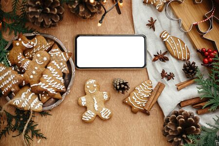 Phone with empty screen and festive gingerbread cookies with anise, cinnamon, pine cones, cedar branches on rustic table, flat lay. Space for text.Merry Christmas. Holiday card mockupの写真素材