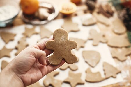 Hand holding raw gingerbread man cookie on background of dough, metal cutters and anise, ginger,...