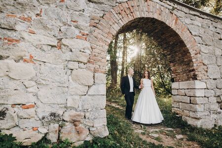 Gorgeous bride and groom embracing in evening sunlight near old castle in park. Romantic moment. Stylish wedding couple gently hugging and walkingの写真素材