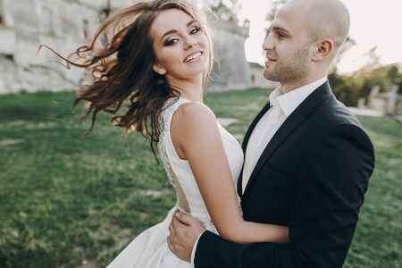 Gorgeous bride and groom having fun and dancing in evening sunlight near old castle in park. Stylish wedding couple gently embracing  and smiling. Romantic momentの写真素材