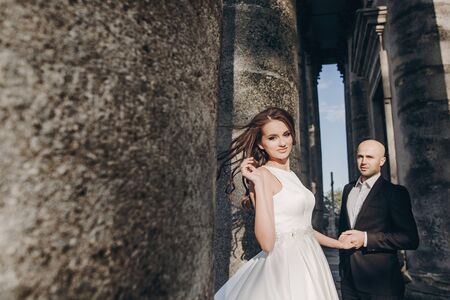 Gorgeous wedding couple embracing in sunlight near old castle in beautiful park. Stylish beautiful bride and groom gently hugging on background of ancient building and natureの写真素材