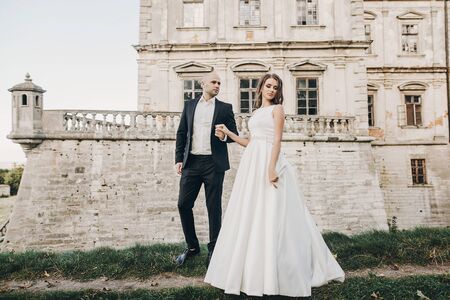 Gorgeous wedding couple walking in sunlight near old castle in beautiful park. Stylish beautiful bride and groom posing on background of ancient building and natureの写真素材