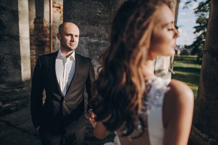 Gorgeous bride and groom posing in sunlight near old castle in park. Stylish wedding couple gently embracing and looking at each other. Romantic momentの写真素材