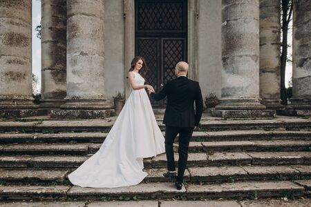 Gorgeous wedding couple walking near old castle in sunny beautiful park. Stylish beautiful bride and groom posing on background of ancient columns and natureの写真素材