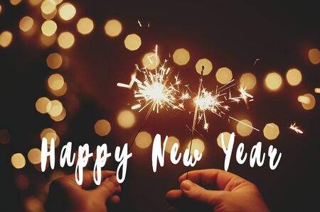 Happy New Year text sign on glowing sparkler in hands on background of golden christmas tree lights in dark festive room. Fireworks burning. Seasons greeting cardの写真素材