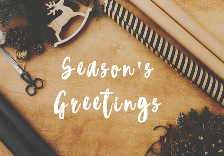 Season's greetings text sign on christmas rustic flat lay, stylish wrapping paper, wooden reindeer, pine branches and cones,  thread on rural wooden table. Season's greeting cardの写真素材