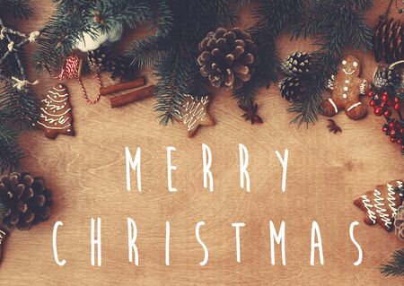 Merry Christmas text sign on stylish rustic frame of pine branches, cones, gingerbread cookies,thread, cinnamon, anise on rural wooden table. Season's greeting card. Flat layの写真素材