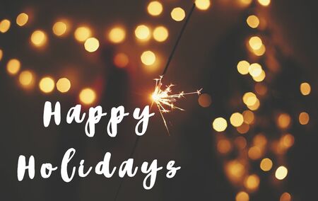 Happy Holidays text sign on glowing sparkler in hand on background of golden christmas tree lights in dark festive room. Fireworks burning. Seasons greeting cardの写真素材