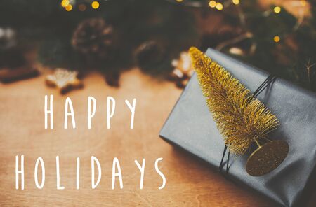 Happy Holidays text sign on stylish black christmas gift with golden tree decoration and pine branches, lights, cones, gingerbread cookies,  on rural wooden table. Season's greeting cardの写真素材