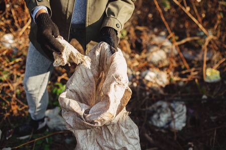 Volunteer picking up dirty plastic  in park. Woman hand in glove picking up trash, collecting garbage in bag. Cleaning up nature from single use plasticの写真素材