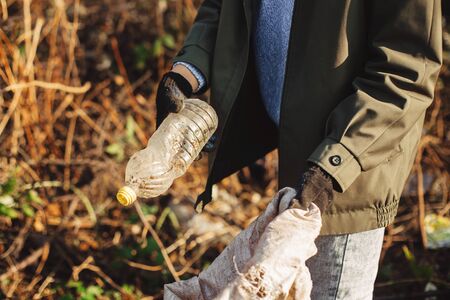 Volunteer picking up dirty plastic bottles in park. Woman hand in glove picking up trash, collecting garbage in bag. Cleaning up nature from single use plasticの写真素材