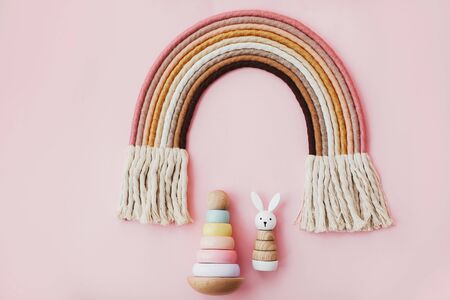 Stylish wooden toys for child on pink background, flat lay. Modern colorful wooden pyramid with rings, wooden bunny and macrame rainbow. Eco friendly plastic free toys for toddlerの写真素材