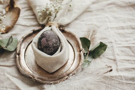 Rustic Easter table decorations. Stylish Easter brunch table setting with egg in easter bunny napkin. Modern natural dyed marble egg on napkin with bunny ears, flowers on vintage plate.の写真素材