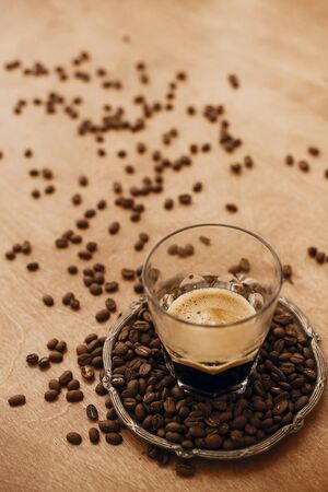 Hot espresso with foam in glass cup on vintage plate with aromatic roasted coffee beans on wooden table. Copy space. Alternative Coffee brewing concept.の写真素材