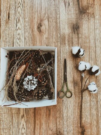Stylish  box with pine cones, herbs,berries, and scissors, cotton on wooden floor flat lay. Making rustic christmas wreath. Preparation for winter holiday decorの写真素材