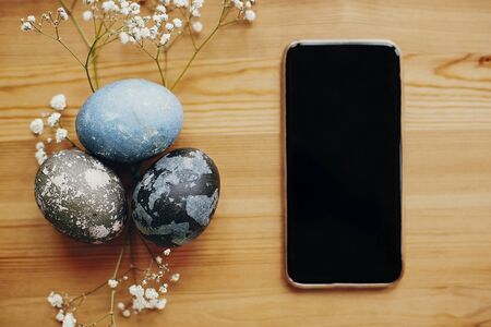 Stylish easter eggs flat lay on rustic table with white spring flowers and phone with empty screen. Natural dyed easter eggs on wooden background with smartphone. Space for textの写真素材