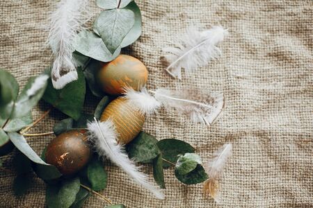 Stylish easter eggs in rustic nest with feathers on rustic table. Natural dyed green easter eggs with eucalyptus branch, spring flowers on rural textile background. Flat layの写真素材