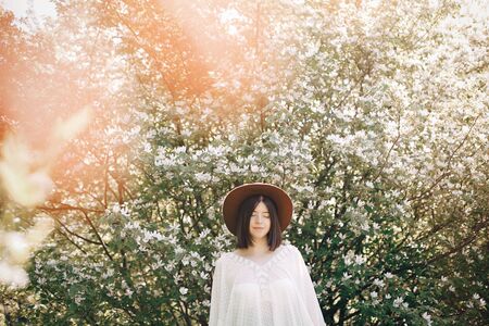 Stylish boho woman in hat posing in blooming tree with white flowers in sunny spring park. Calm...