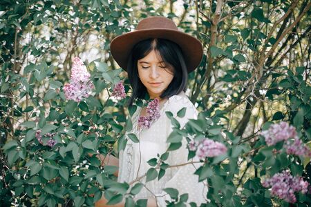 Stylish boho woman in hat posing at lilac flowers in sunny spring park. Calm portrait of beautiful hipster girl standing in purple lilac bush in spring garden.の写真素材