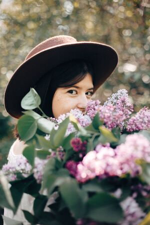 Sensual portrait of beautiful boho woman in hat holding lilac flowers bouquets in sunny spring park. Stylish calm hipster girl with purple lilac posing in spring garden. Copy spaceの写真素材