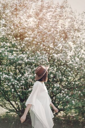 Blurred image of happy boho woman in hat dancing in sunny light near white blooming tree in spring park. Stylish hipster girl enjoying spring. Atmospheric moment of happinessの写真素材