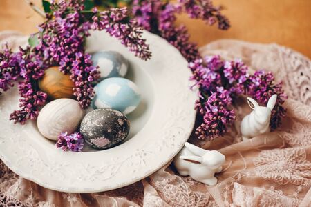 Happy Easter. Stylish Easter eggs on vintage plate, white bunnies and lilac flowers on fabric on...