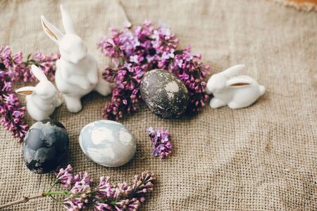 Modern Easter eggs, white bunnies and lilac flowers on rustic fabric. Stylish holiday table decor....