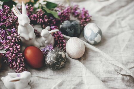 Modern Easter eggs, white bunnies and lilac flowers on linen rural fabric. Stylish holiday table...