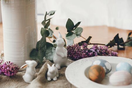 Stylish Easter eggs on vintage plate, white bunny rabbits and lilac flowers on rustic fabric on...