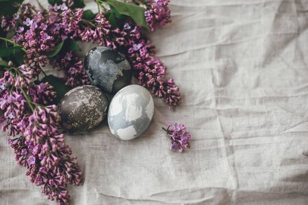 Modern Easter eggs on rustic linen fabric and purple lilac flowers. Rural still life. Happy Easter....