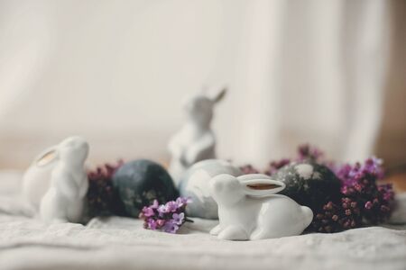 Happy Easter. Modern Easter eggs, white bunnies and lilac flowers on linen rustic fabric. Stylish...