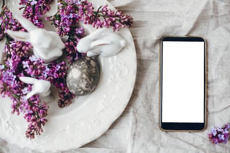 Phone with empty blank screen and stylish easter white bunnies, modern easter egg on vintage plate and lilac flowers on linen rural fabric, flat lay. Space for text. Happy Easter mockupの写真素材