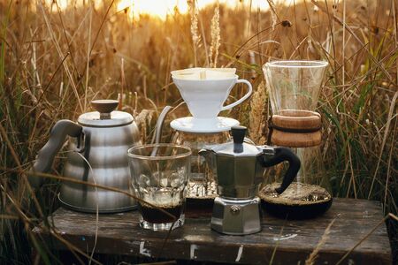 Alternative coffee brewing outdoors in travel. Steel kettle, hot coffee in cup, coffee dripper,  geyser maker, glass flask with filter on background of sunny warm light in rural herbs.の写真素材