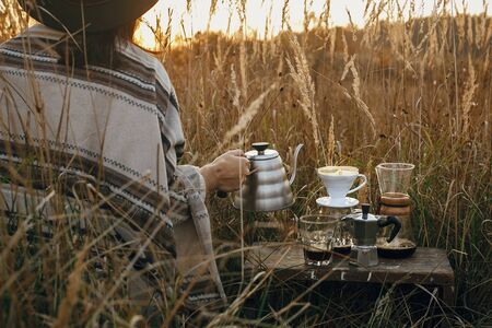 Alternative coffee brewing outdoors in travel. Hipster woman holding hot kettle and coffee cup, glass flask, geyser maker on background of sunny warm light in rural herbs. Back view. Authentic momentの写真素材