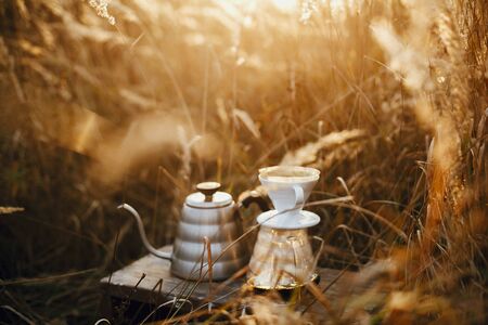 Steel kettle and glass flask dripper with filter on background of rural countryside herbs in sunset. Alternative coffee brewing outdoors. Atmospheric rustic moment. Space for textの写真素材