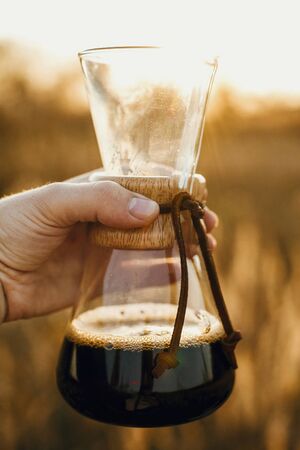 Alternative coffee brewing outdoors in travel. Hand holding hot coffee in glass flask on background in sunny warm light in rural herbs.Atmospheric rustic tranquil moment.の写真素材